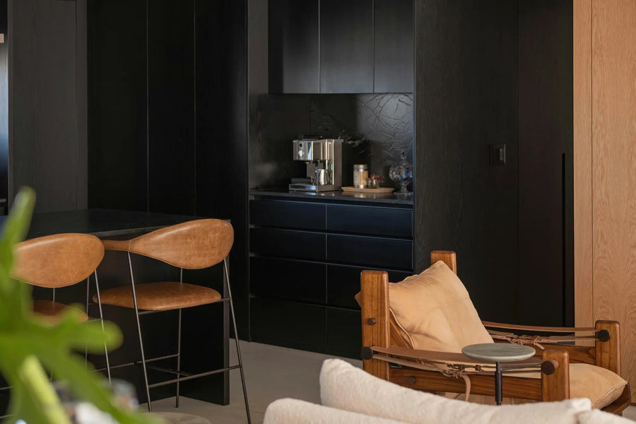 Tummasävyinen moderni keittiö, jossa nahkaiset baarituolit ja mustat kaapistot luovat tyylikkään kontrastin. Esimerkki rohkeista sävyistä ja luonnonmateriaaleista vuoden 2026 keittiötrendeissä.