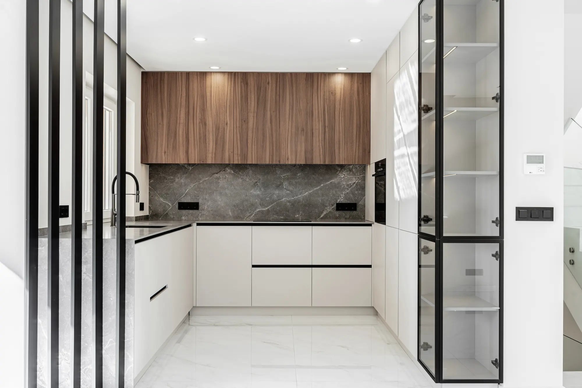 : Moderni keittiö, jossa työtaso ja nostettu välitila harmaasta kivikuosisesta materiaalista ja tummanruskeat yläkaapit puuviilua. Etualalla lasiovinen vitriinikaappi. Esimerkki ajattomista materiaaleista keittiössä.