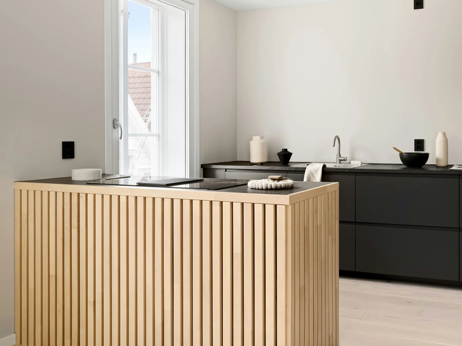 Moderni keittiö, jossa on vaalea puinen keittiösaareke, mustat kaapistot ja harmaa työtaso, sekä ikkuna, josta tulee luonnonvaloa.