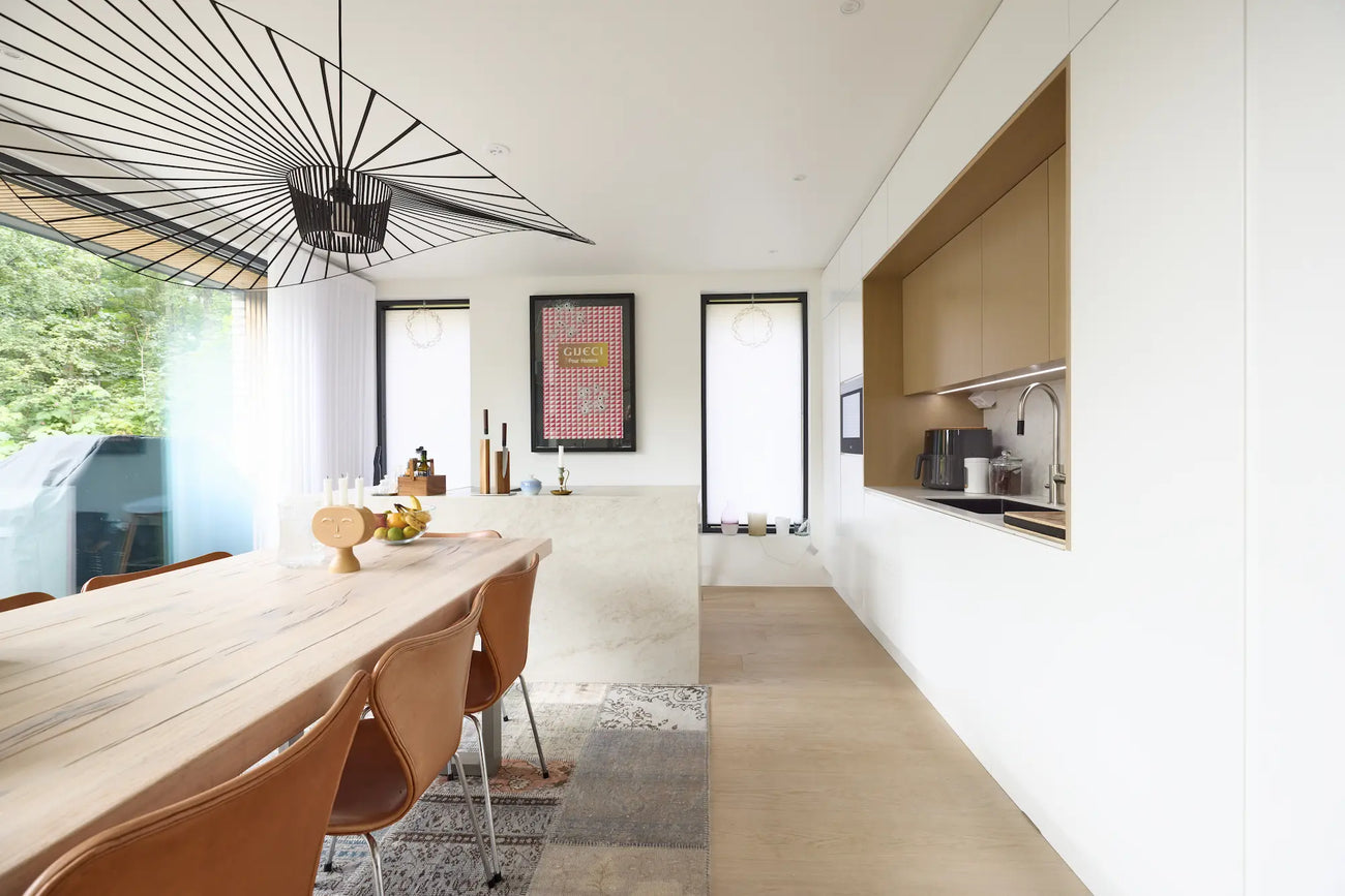 Vaalea ja harmoninen keittiö- ja ruokailutila, jossa yhdistyvät luonnonmateriaalit, tammiviiluiset kaapistot ja suuri ruokapöytä nahkatuoleineen. Musta design-valaisin ja isot ikkunat tuovat tilaan ilmettä ja luonnonvaloa.